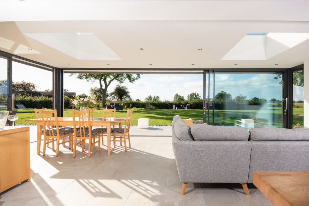 Modern living room with a gray sofa and wooden dining table. Large sliding glass doors by HBD Systems open to a lush garden view, filling the space with natural light from the glazed extension.