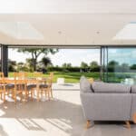 Thumbnail of http://Modern%20living%20room%20with%20a%20gray%20sofa%20and%20wooden%20dining%20table.%20Large%20sliding%20glass%20doors%20by%20HBD%20Systems%20open%20to%20a%20lush%20garden%20view,%20filling%20the%20space%20with%20natural%20light%20from%20the%20glazed%20extension.