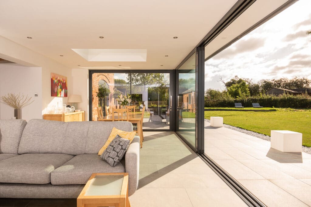 A modern living room features a gray sofa and wooden dining table, with large sliding doors from HBD Systems leading to a grassy backyard under a partly cloudy sky, providing an elegant glazed extension to the space.