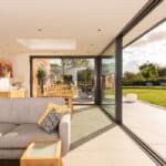 Thumbnail of http://A%20modern%20living%20room%20features%20a%20gray%20sofa%20and%20wooden%20dining%20table,%20with%20large%20sliding%20doors%20from%20HBD%20Systems%20leading%20to%20a%20grassy%20backyard%20under%20a%20partly%20cloudy%20sky,%20providing%20an%20elegant%20glazed%20extension%20to%20the%20space.