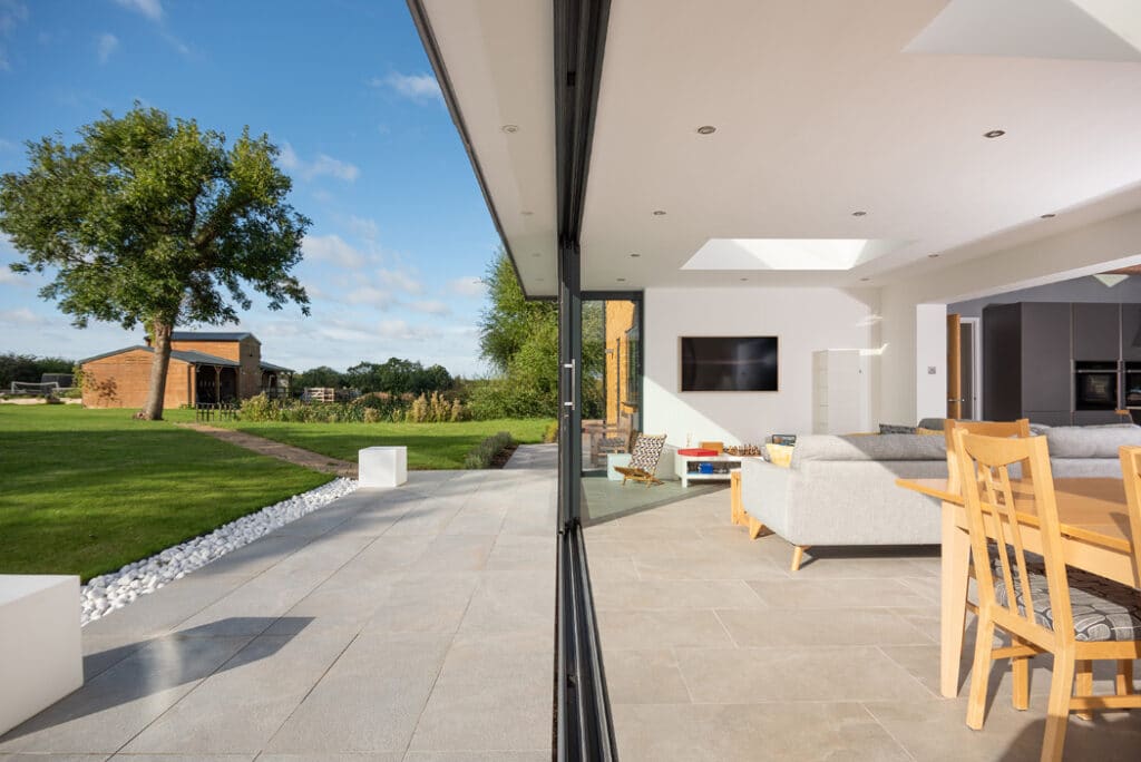 Modern living area with sliding doors opens to reveal a landscaped garden featuring an extension by HBD Systems. Large windows frame a serene view of the tree and outbuilding. Inside, enjoy the comfort of a sofa, dining table, and wall-mounted TV.