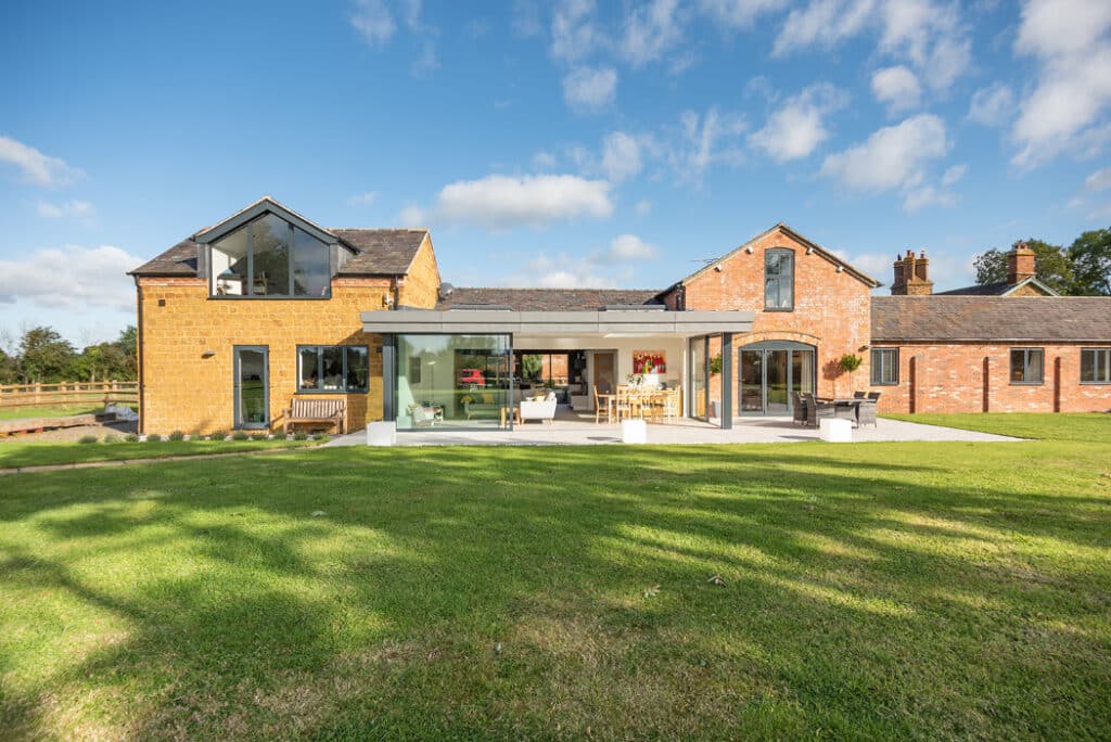 Spacious modern house with large glass windows and a red brick exterior, featuring HBD Systems' sliding doors and a stylish glazed extension. It's surrounded by a green lawn under a blue sky with clouds.