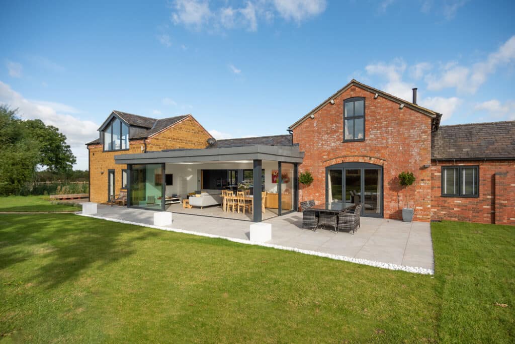 A modern house exterior featuring a blend of brick and glass, with a spacious patio and outdoor seating. The adjacent lawn is accessible through sleek sliding doors by HBD Systems, seamlessly merging indoor and outdoor spaces under a clear sky.