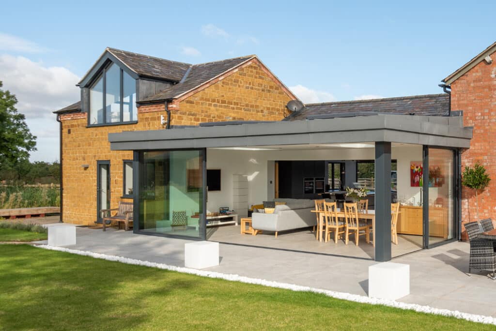 Modern house with large glass windows and a stunning HBD Systems glazed extension, featuring sliding doors that open onto an outdoor patio with seating. Enjoy the well-manicured lawn under a clear sky.