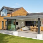 Thumbnail of http://Modern%20house%20with%20large%20glass%20windows%20and%20a%20stunning%20HBD%20Systems%20glazed%20extension,%20featuring%20sliding%20doors%20that%20open%20onto%20an%20outdoor%20patio%20with%20seating.%20Enjoy%20the%20well-manicured%20lawn%20under%20a%20clear%20sky.