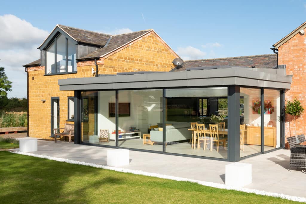 Modern house featuring a large HBD Systems glazed extension with sliding doors, brick exterior, and a patio. Inside, it boasts a dining table and seating area surrounded by well-maintained grass.