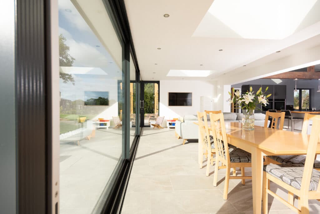 Modern living room with HBD Systems' glazed sliding doors, a sleek wooden dining table, chairs, and light gray sofas. The wall-mounted TV sits elegantly as natural light floods in from skylights and the window, creating an inviting ambience throughout the extension.