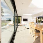 Thumbnail of http://Modern%20living%20room%20with%20HBD%20Systems'%20glazed%20sliding%20doors,%20a%20sleek%20wooden%20dining%20table,%20chairs,%20and%20light%20gray%20sofas.%20The%20wall-mounted%20TV%20sits%20elegantly%20as%20natural%20light%20floods%20in%20from%20skylights%20and%20the%20window,%20creating%20an%20inviting%20ambience%20throughout%20the%20extension.