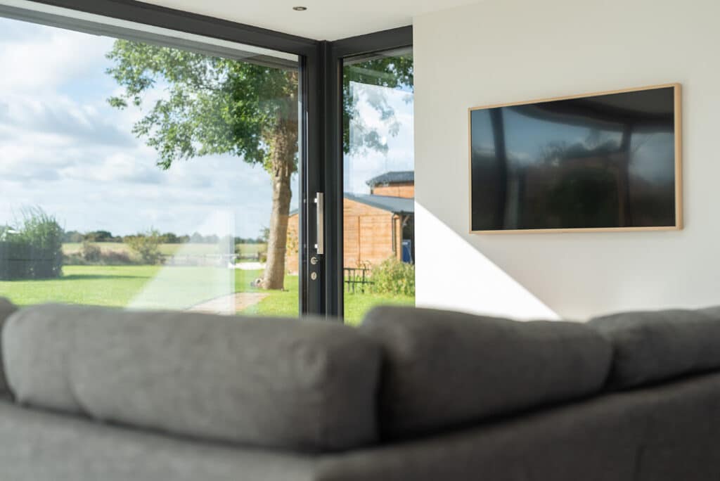 The living room features a gray sofa and large windows, complemented by a mounted flat-screen TV on the wall. Sliding doors by HBD Systems open to reveal a lush green lawn, seamlessly blending indoor comfort with outdoor beauty.
