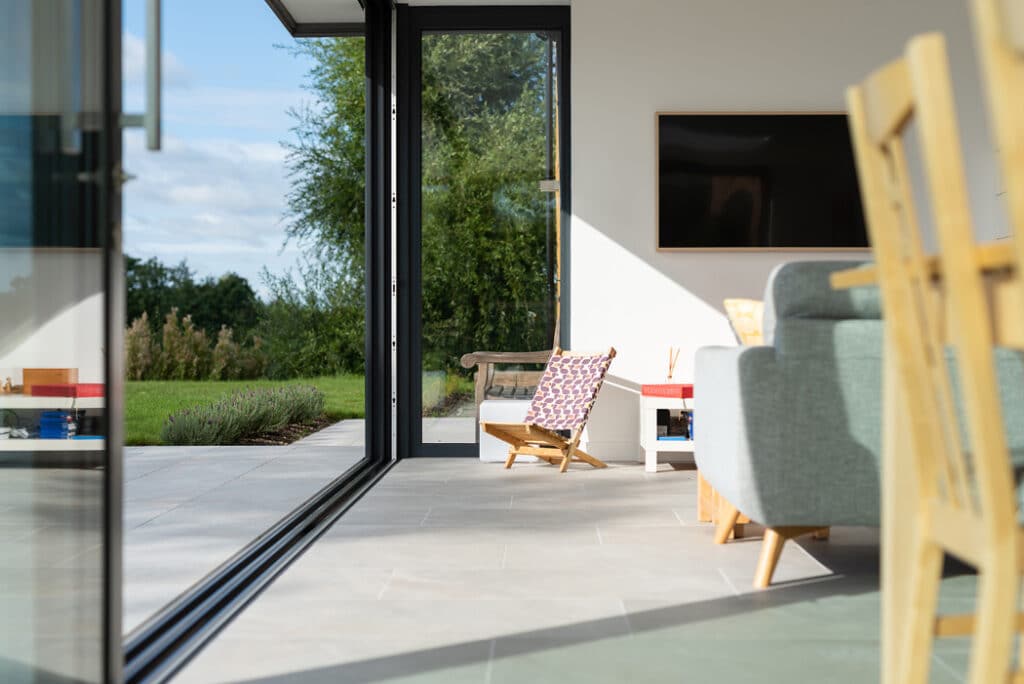 Modern living room with sliding glass doors by HBD Systems that open to a patio; a chair with a patterned pillow basks in the sunlight from the glazed extension.