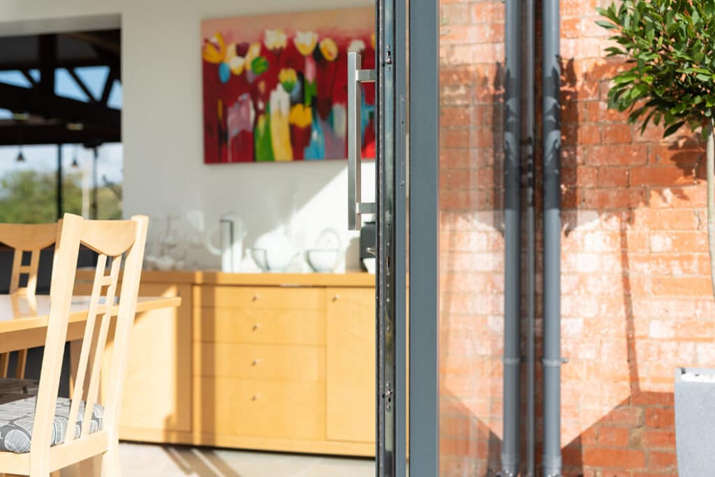 An open sliding glass door reveals a dining area with a wooden table and chairs. A colorful flower painting hangs on the wall above a sideboard, while sunlight from the HBD Systems glazed extension illuminates the space.