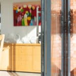 Thumbnail of http://An%20open%20sliding%20glass%20door%20reveals%20a%20dining%20area%20with%20a%20wooden%20table%20and%20chairs.%20A%20colorful%20flower%20painting%20hangs%20on%20the%20wall%20above%20a%20sideboard,%20while%20sunlight%20from%20the%20HBD%20Systems%20glazed%20extension%20illuminates%20the%20space.