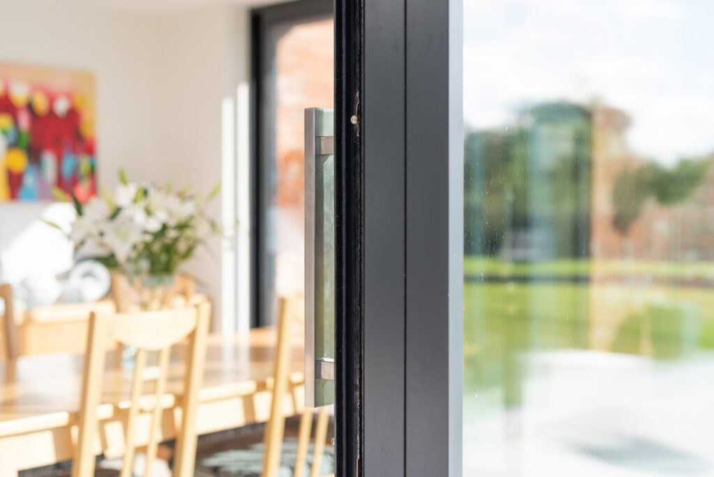 Close-up of a slightly open sliding glass door by HBD Systems, offering a glimpse into a bright dining area. Inside, there's a table, chairs, a colorful painting, and a vase with flowers. The space beautifully connects to the garden outside through the glazed extension.