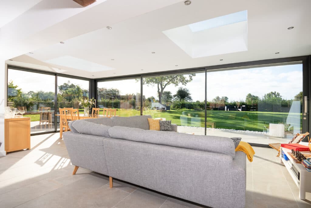 The modern living room features a grey sofa and large windows with a stunning view of the green garden. Skylights and sliding doors by HBD Systems ensure ample natural light floods the space. A dining table is charmingly visible in the glazed extension, blending indoor and outdoor serenity.