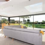 Thumbnail of http://The%20modern%20living%20room%20features%20a%20grey%20sofa%20and%20large%20windows%20with%20a%20stunning%20view%20of%20the%20green%20garden.%20Skylights%20and%20sliding%20doors%20by%20HBD%20Systems%20ensure%20ample%20natural%20light%20floods%20the%20space.%20A%20dining%20table%20is%20charmingly%20visible%20in%20the%20glazed%20extension,%20blending%20indoor%20and%20outdoor%20serenity.