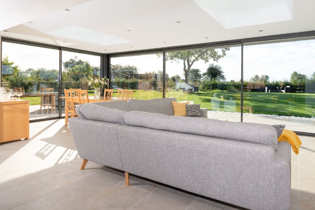 The spacious living room features a gray sofa, large windows, and sleek sliding doors from HBD Systems, offering an open view of the garden and dining area in a stunning glazed extension.