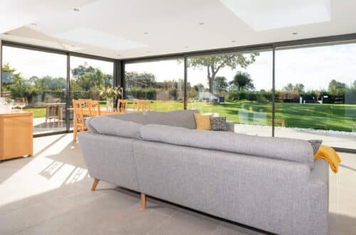 The spacious living room features a gray sofa, large windows, and sleek sliding doors from HBD Systems, offering an open view of the garden and dining area in a stunning glazed extension.