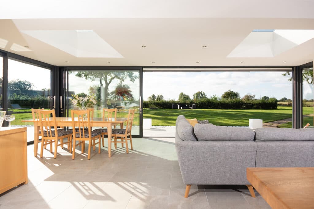Modern living room featuring a gray sofa, wooden table, and chairs. Large windows and sliding doors by HBD Systems lead to a glazed extension, offering views of the green lawn and trees. Skylights brighten the space beautifully.