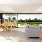 Thumbnail of http://Modern%20living%20room%20featuring%20a%20gray%20sofa,%20wooden%20table,%20and%20chairs.%20Large%20windows%20and%20sliding%20doors%20by%20HBD%20Systems%20lead%20to%20a%20glazed%20extension,%20offering%20views%20of%20the%20green%20lawn%20and%20trees.%20Skylights%20brighten%20the%20space%20beautifully.