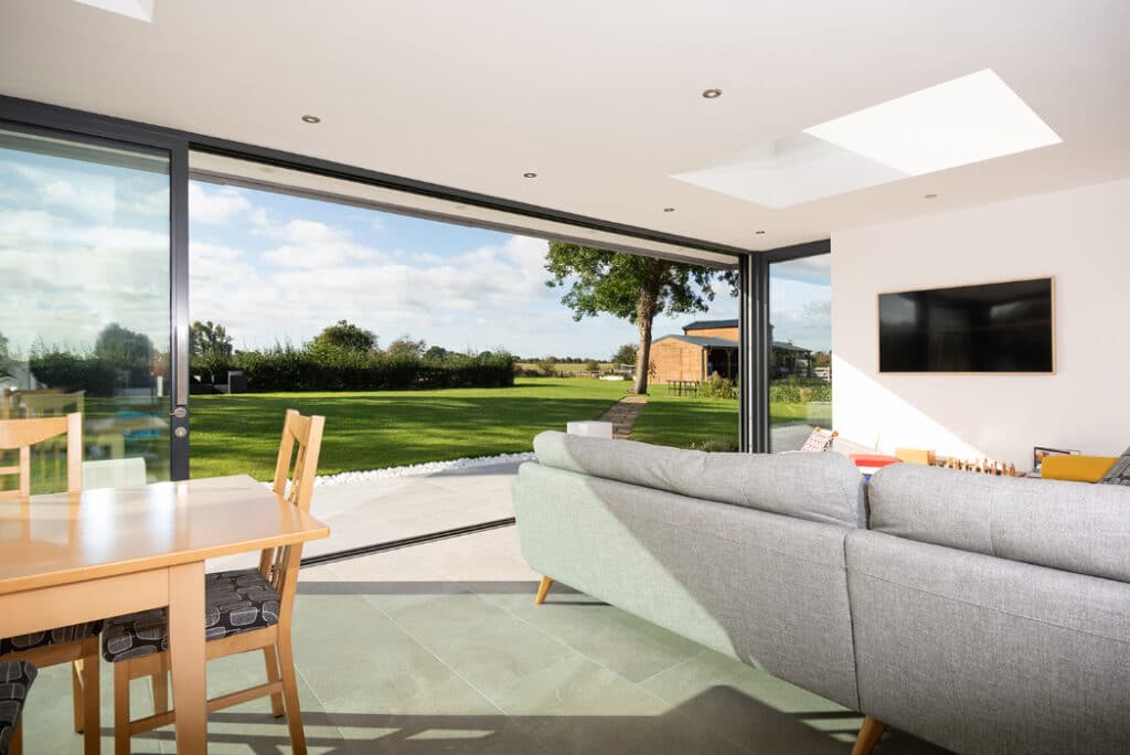 Modern living room with sliding doors by HBD Systems, overlooking a sunny garden with trees. This glazed extension enhances the space, featuring a gray sofa, wooden dining table, and wall-mounted TV.