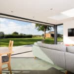 Thumbnail of http://Modern%20living%20room%20with%20sliding%20doors%20by%20HBD%20Systems,%20overlooking%20a%20sunny%20garden%20with%20trees.%20This%20glazed%20extension%20enhances%20the%20space,%20featuring%20a%20gray%20sofa,%20wooden%20dining%20table,%20and%20wall-mounted%20TV.