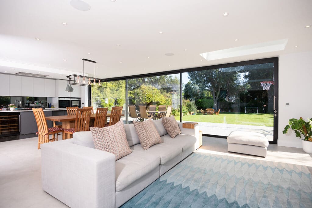 Modern open-plan living space with a sofa, dining area, and large sliding glass doors by HBD Systems leading to a garden with a basketball hoop. The room features a light color scheme and contemporary decor.
