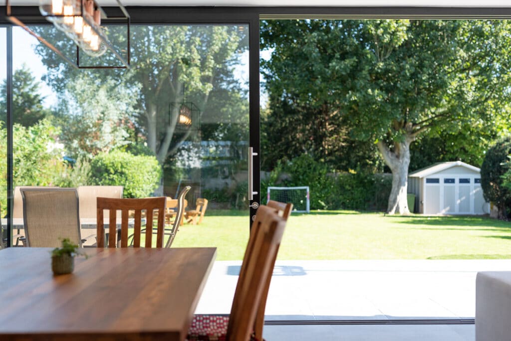 A modern dining area with elegant wooden furniture opens to a backyard through sleek sliding doors by HBD Systems. The charming backyard features a large tree, a cozy shed, and a goalpost, creating an inviting space for relaxation and play.