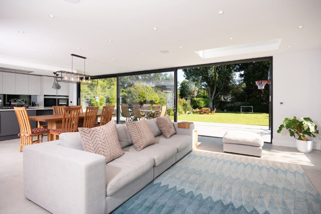 Modern open-plan living room featuring a gray sofa, patterned cushions, and a wooden dining set. Large sliding doors by HBD Systems lead to a garden complete with a basketball hoop and soccer goal.