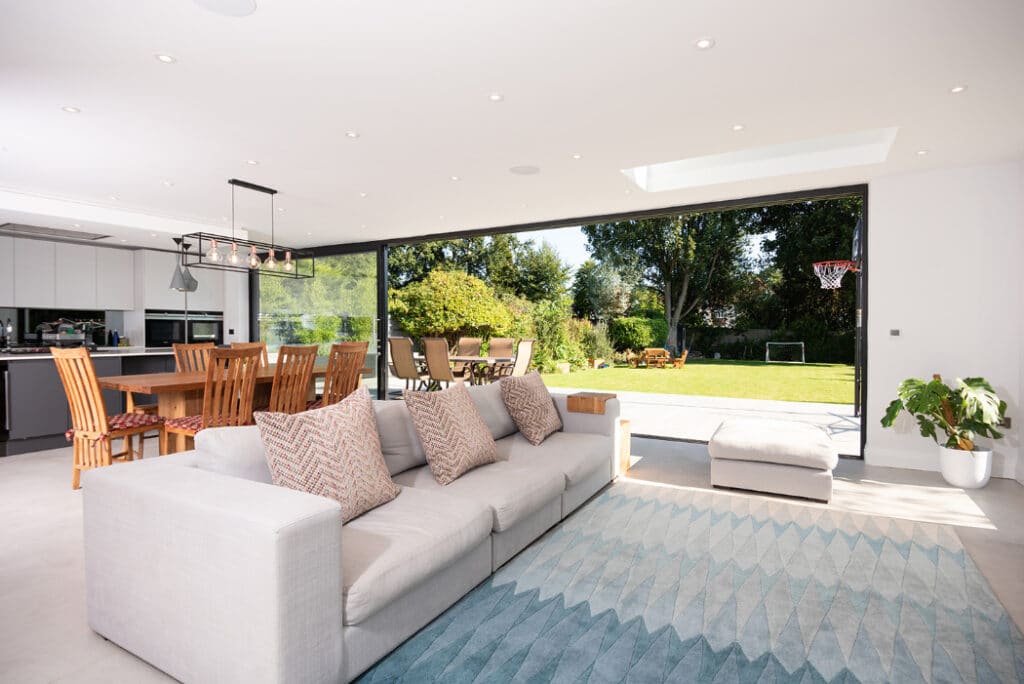 Spacious living room with a gray sofa and patterned rug, featuring HBD Systems sliding doors that open to a garden with trees and a basketball hoop. A dining table and kitchen area are visible in the background.