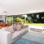 Thumbnail of http://Spacious%20living%20room%20with%20a%20gray%20sofa%20and%20patterned%20rug,%20featuring%20HBD%20Systems%20sliding%20doors%20that%20open%20to%20a%20garden%20with%20trees%20and%20a%20basketball%20hoop.%20A%20dining%20table%20and%20kitchen%20area%20are%20visible%20in%20the%20background.