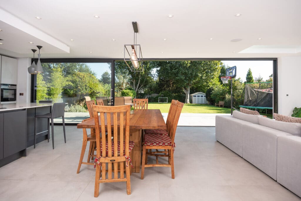 Modern open-plan kitchen and dining area featuring a wooden table and chairs, light gray tiles, and floor-to-ceiling sliding doors by HBD Systems. These doors lead to a garden equipped with a patio, trampoline, and basketball hoop.