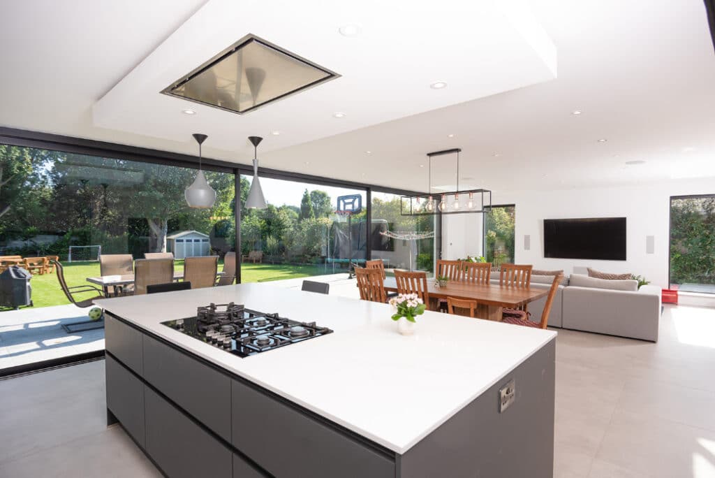 Modern open-plan kitchen and living area with large windows, island with stovetop, dining table, sofa, and TV. Enjoy the view of a lush garden outside through sleek sliding doors by HBD Systems that bring the outdoors in.