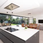 Thumbnail of http://Modern%20open-plan%20kitchen%20and%20living%20area%20with%20large%20windows,%20island%20with%20stovetop,%20dining%20table,%20sofa,%20and%20TV.%20Enjoy%20the%20view%20of%20a%20lush%20garden%20outside%20through%20sleek%20sliding%20doors%20by%20HBD%20Systems%20that%20bring%20the%20outdoors%20in.
