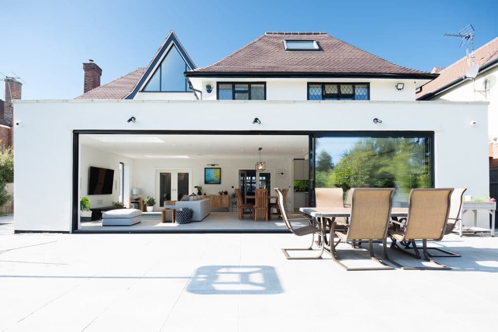 The modern house features large glass sliding doors by HBD Systems, opening to a spacious patio. An outdoor dining set and cushioned seating grace the white-tiled terrace under the clear blue sky.