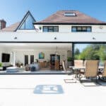 Thumbnail of http://The%20modern%20house%20features%20large%20glass%20sliding%20doors%20by%20HBD%20Systems,%20opening%20to%20a%20spacious%20patio.%20An%20outdoor%20dining%20set%20and%20cushioned%20seating%20grace%20the%20white-tiled%20terrace%20under%20the%20clear%20blue%20sky.