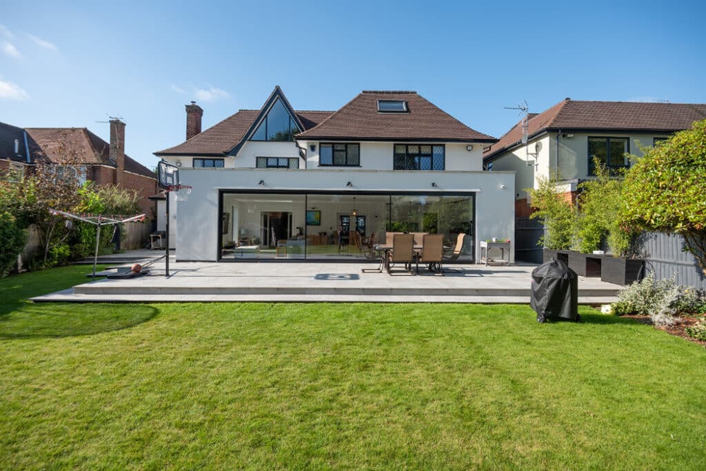 Rear view of a modern house featuring HBD Systems' sleek sliding doors, a patio area with dining set, barbecue, and a spacious green lawn under a clear blue sky.