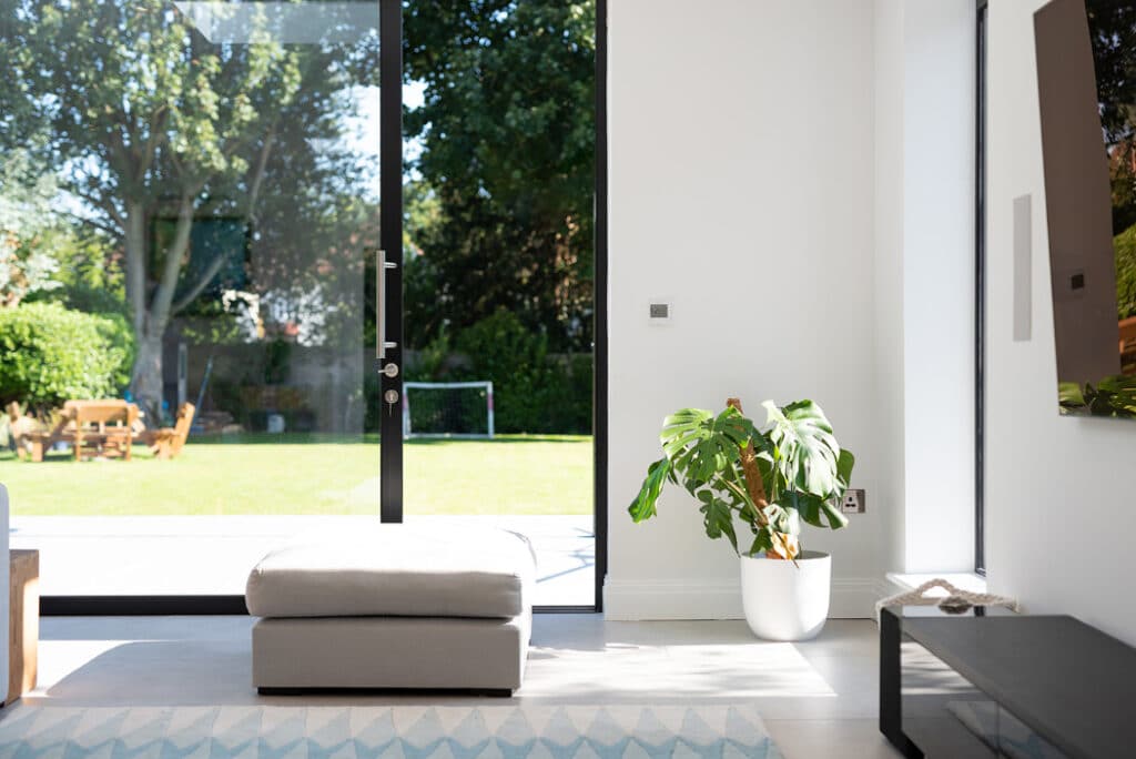 Bright room with large sliding doors by HBD Systems leading to a garden. A white potted plant and a gray ottoman sit near the door. Outside, a children's play area is visible on the lawn.