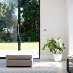 Thumbnail of http://Bright%20room%20with%20large%20sliding%20doors%20by%20HBD%20Systems%20leading%20to%20a%20garden.%20A%20white%20potted%20plant%20and%20a%20gray%20ottoman%20sit%20near%20the%20door.%20Outside,%20a%20children's%20play%20area%20is%20visible%20on%20the%20lawn.