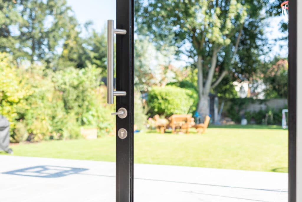 Sliding doors from HBD Systems open to a sunny backyard with a lush green lawn, trees, and inviting patio furniture.