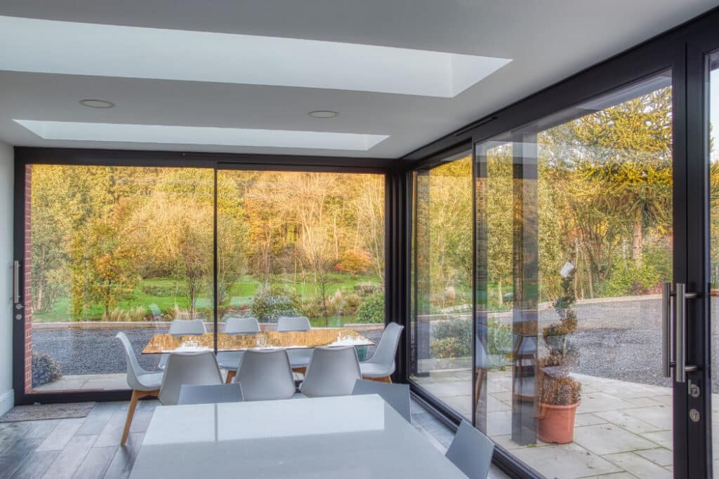 Modern dining area with glass walls and ceiling, overlooking a patio and garden with trees.