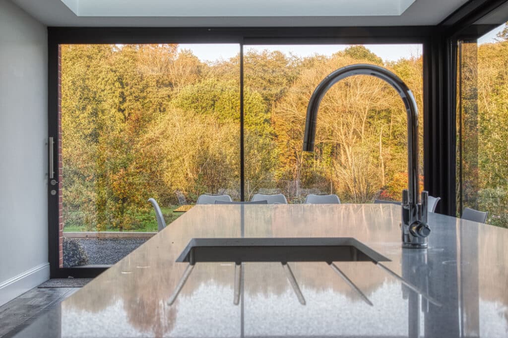 Modern kitchen with a sleek island and faucet, featuring large glass doors opening to a scenic view of autumn trees.