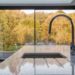 Thumbnail of http://Modern%20kitchen%20with%20a%20sleek%20island%20and%20faucet,%20featuring%20large%20glass%20doors%20opening%20to%20a%20scenic%20view%20of%20autumn%20trees.