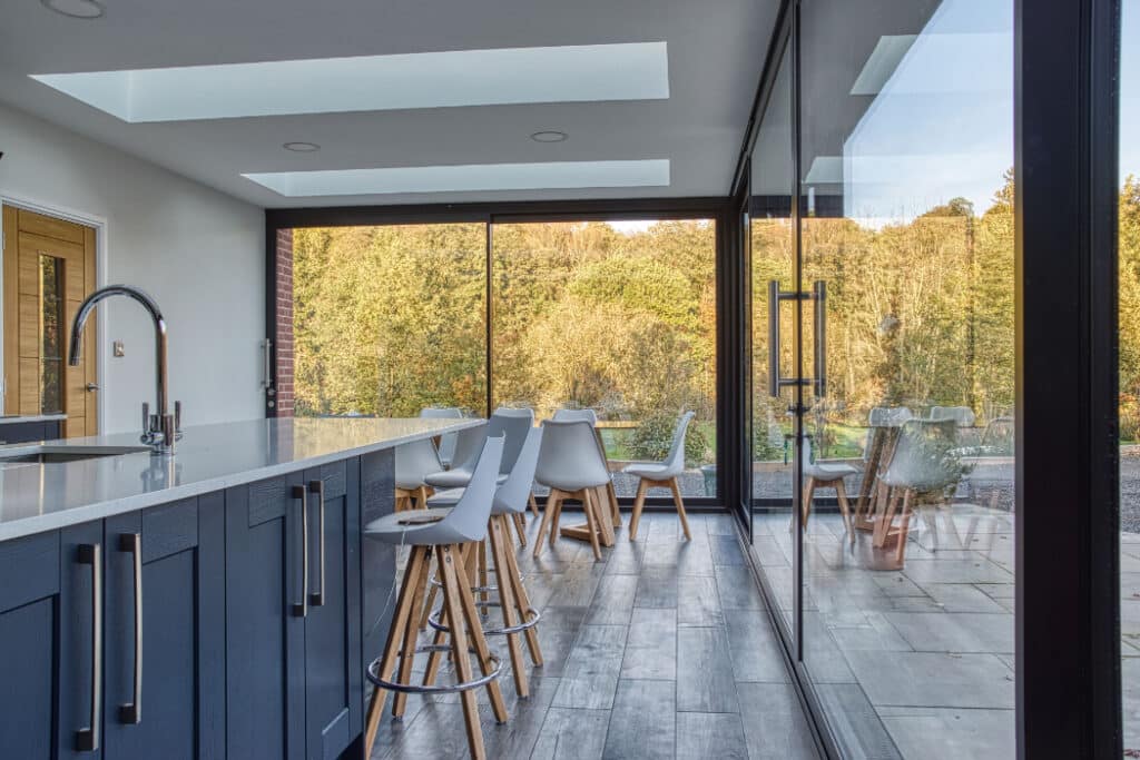 Modern kitchen interior with a long island, bar stools, and floor-to-ceiling windows showing a view of trees outside.