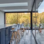 Thumbnail of http://Modern%20kitchen%20interior%20with%20a%20long%20island,%20bar%20stools,%20and%20floor-to-ceiling%20windows%20showing%20a%20view%20of%20trees%20outside.