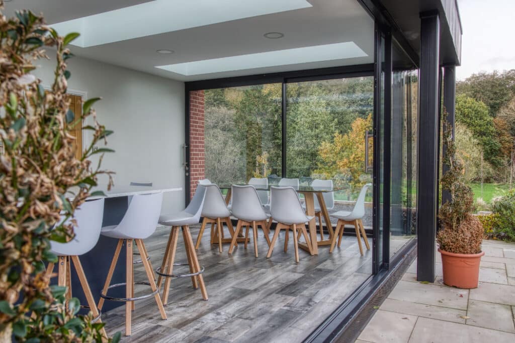 Modern dining area with glass walls, wooden floor, and white chairs around a long table. Outdoor greenery visible through large windows.
