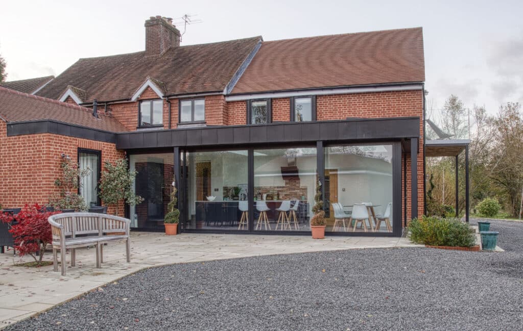A red brick house with a modern glass extension, featuring a patio area with outdoor seating and potted plants.