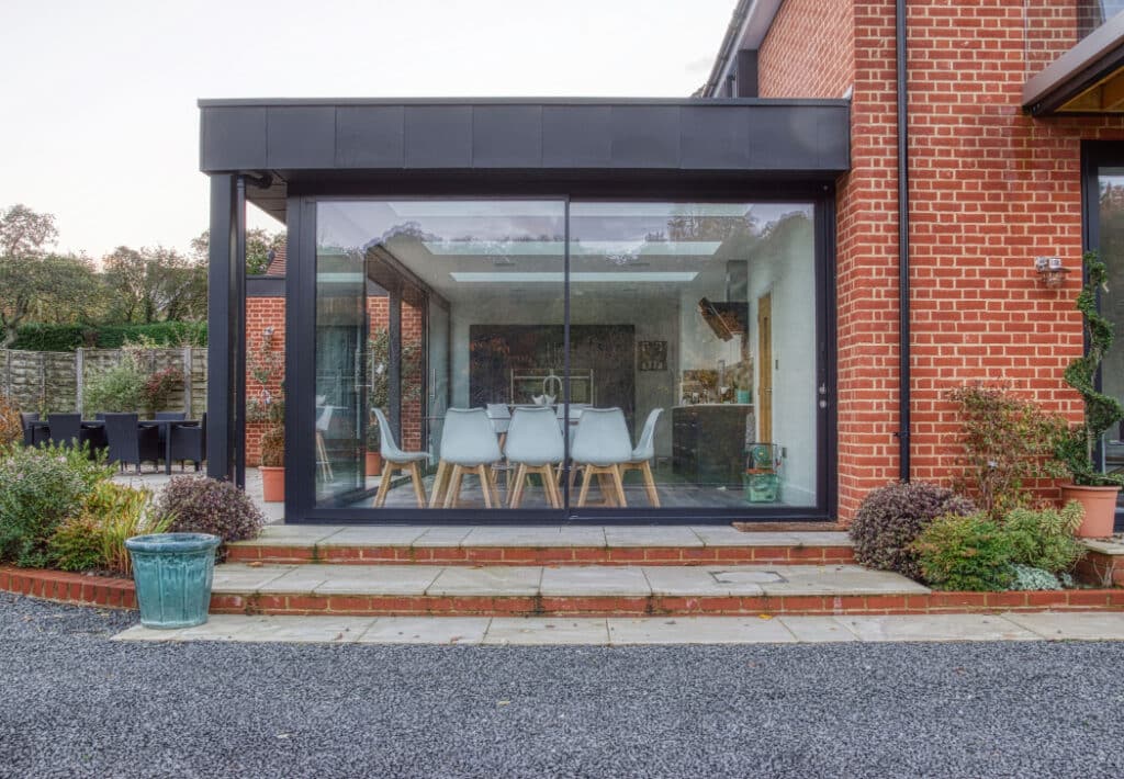 Glass-enclosed dining area with modern white chairs and a table, attached to a red brick building. Patio steps and gravel path are in the foreground.