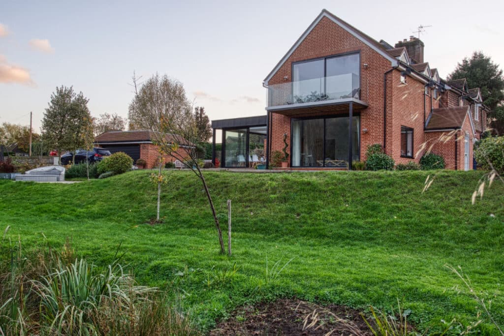 Brick house with large glass windows and balcony, surrounded by grass and trees, under a partly cloudy sky.