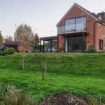 Thumbnail of http://Brick%20house%20with%20large%20glass%20windows%20and%20balcony,%20surrounded%20by%20grass%20and%20trees,%20under%20a%20partly%20cloudy%20sky.