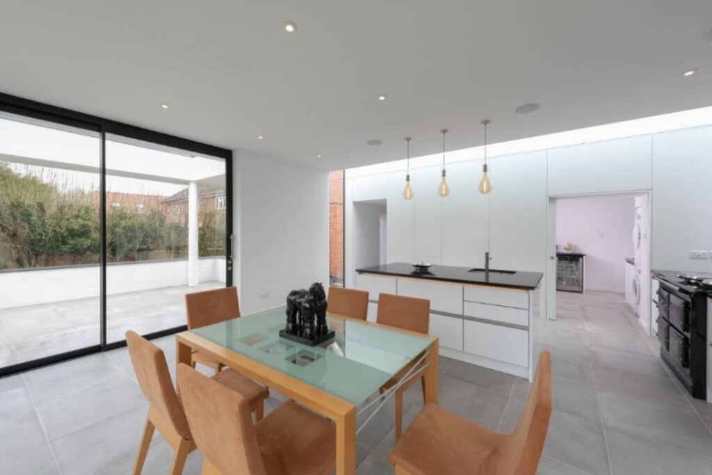 Modern dining area with a glass table and six tan chairs, next to an open kitchen with a black countertop. Large sliding glass doors open to a spacious patio.